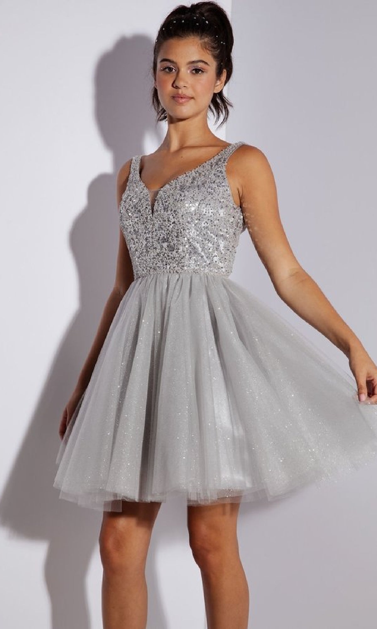 A woman stands against a plain background wearing the Glitter-Bodice Short A-Line Hoco Dress: Eureka 9926, which features a sequin bodice and flared tulle skirt.